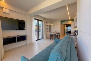 A modern living room with a large flat-screen TV, a sofa, a dining area, and a kitchen.
