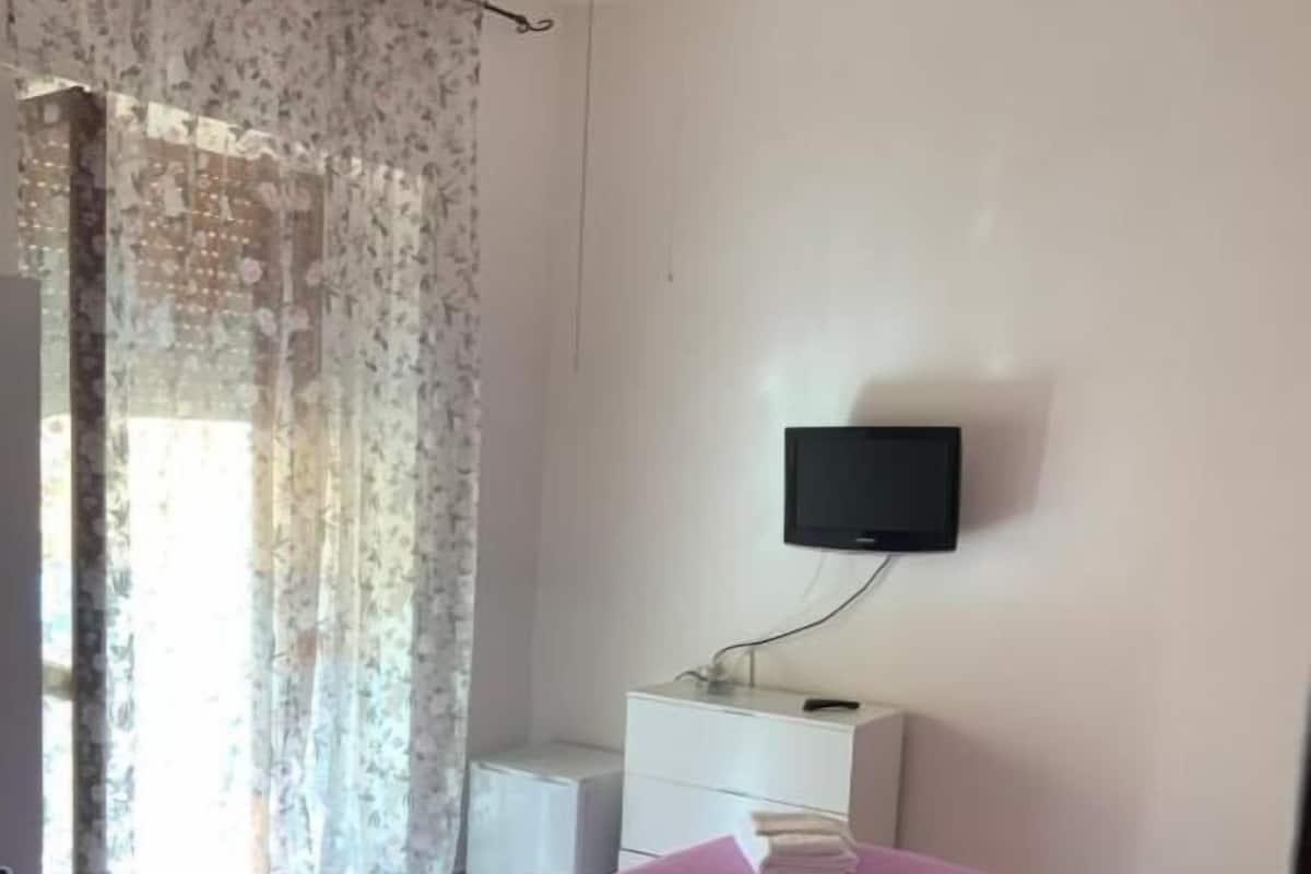 Un dormitorio con una cama, un televisor montado en la pared, una mesita de noche y una ventana con cortinas.
