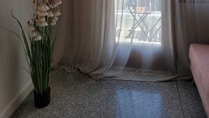 Une pièce dotée d’une grande fenêtre ornée de rideaux légers, d’une plante en pot aux fleurs blanches et offrant une vue sur un balcon aménagé avec du mobilier d’extérieur.