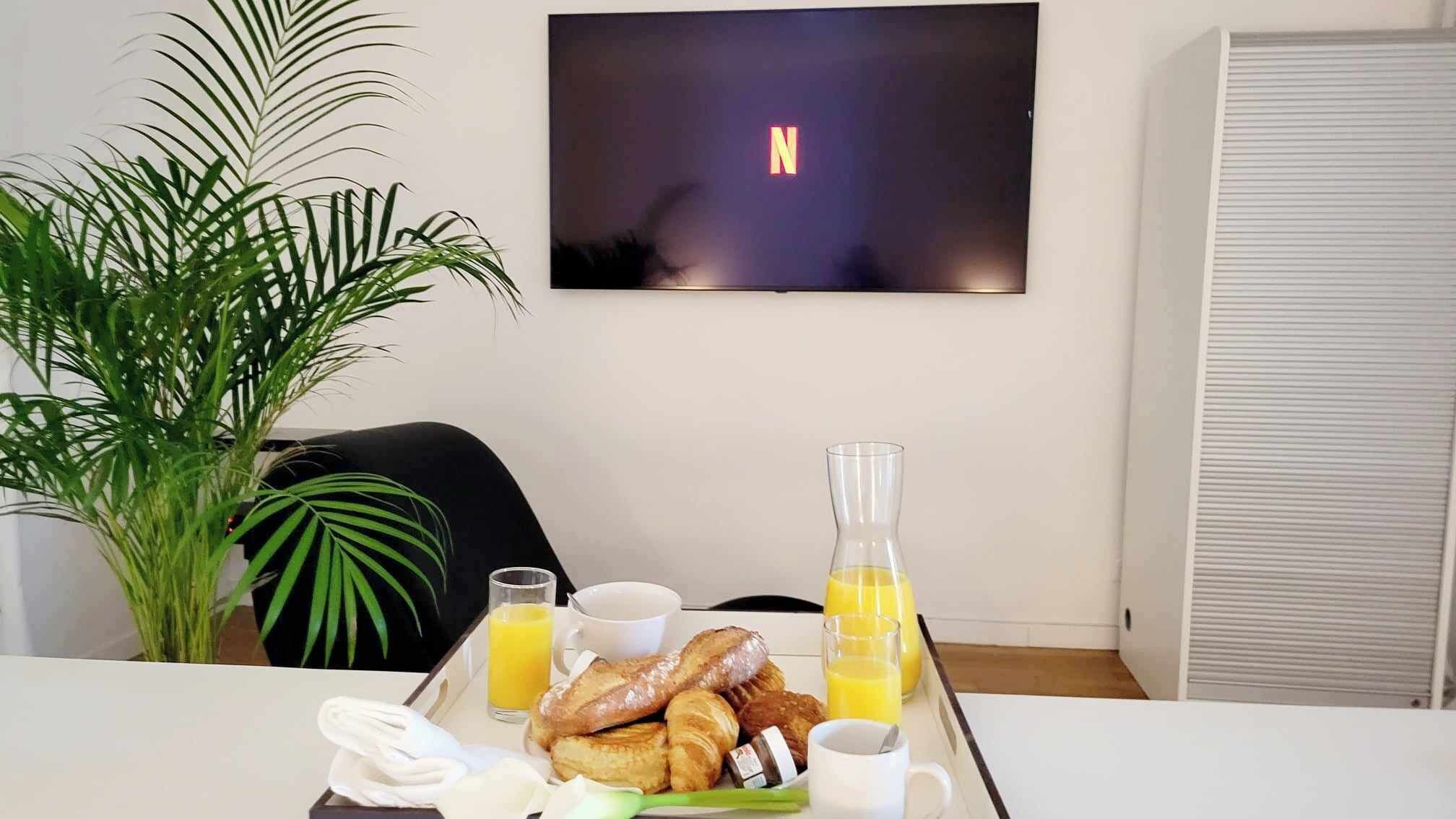 A breakfast tray with croissants, juice, and coffee on a table in a room with a TV displaying the Netflix logo.