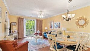 A living room with a dining area, a ceiling fan, and a large window with curtains. - Havens On The Nine 2 Bedroom Condo (North Myrtle Beach)