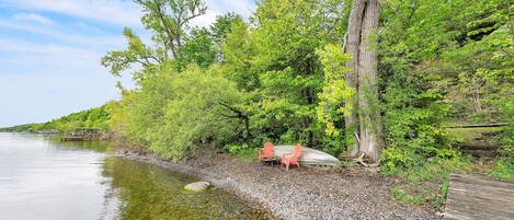 A riverside area with a wooden deck, two red chairs, and a large tree.