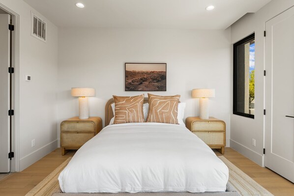 Une chambre à coucher avec un grand lit, deux tables de chevet avec des lampes, un tableau accroché au mur et une fenêtre donnant sur un beau paysage.