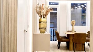 A modern dining area with a white table and brown chairs, a vase with dried flowers, and a view of the city through large windows.