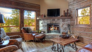 A cozy living room with a stone fireplace, leather furniture, and large windows offering mountain views. - 5BR Ski-In/Ski-Out Cabin, Hot Tub, Fireplace & Mountain Views – Big Sky, MT (Big Sky)