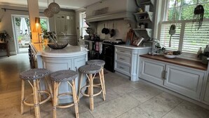 A kitchen with white cabinetry, a central island with stools, hanging pots, and a window with blinds. - Stunning Rectory, 15 guests in Surrey (South Nutfield)