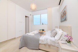A modern bedroom with a bed, bedside table, and a window with a view. - Green Bay Villa 14 in Protaras (Paralimni)