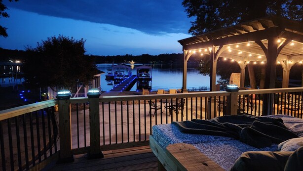 Une terrasse en bois avec un lit, un belvédère illuminé de guirlandes lumineuses, et une vue sur un quai et une maison situés de l’autre côté de l’eau.