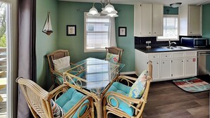 A dining area with a glass-top table surrounded by wicker chairs with teal cushions, a kitchen with white cabinets, and a window with blinds.
