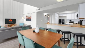 A modern living room with a wooden dining table, green chairs, a grey sofa, and a flat-screen TV.