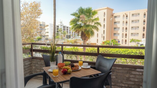 A balcony with a table set for breakfast, including fruits, pastries, and beverages, overlooking a cityscape with buildings and palm trees. - Las Gacelas Playa del Ingles (San Bartolome de Tirajana)