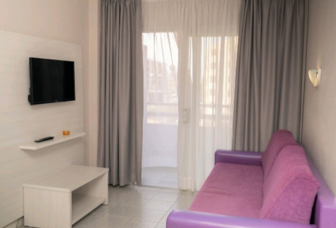Una sala de estar moderna con un sofá morado, un televisor de pantalla plana montado en la pared, un pequeño banco blanco y un puerta corrediza de vidrio con cortinas.