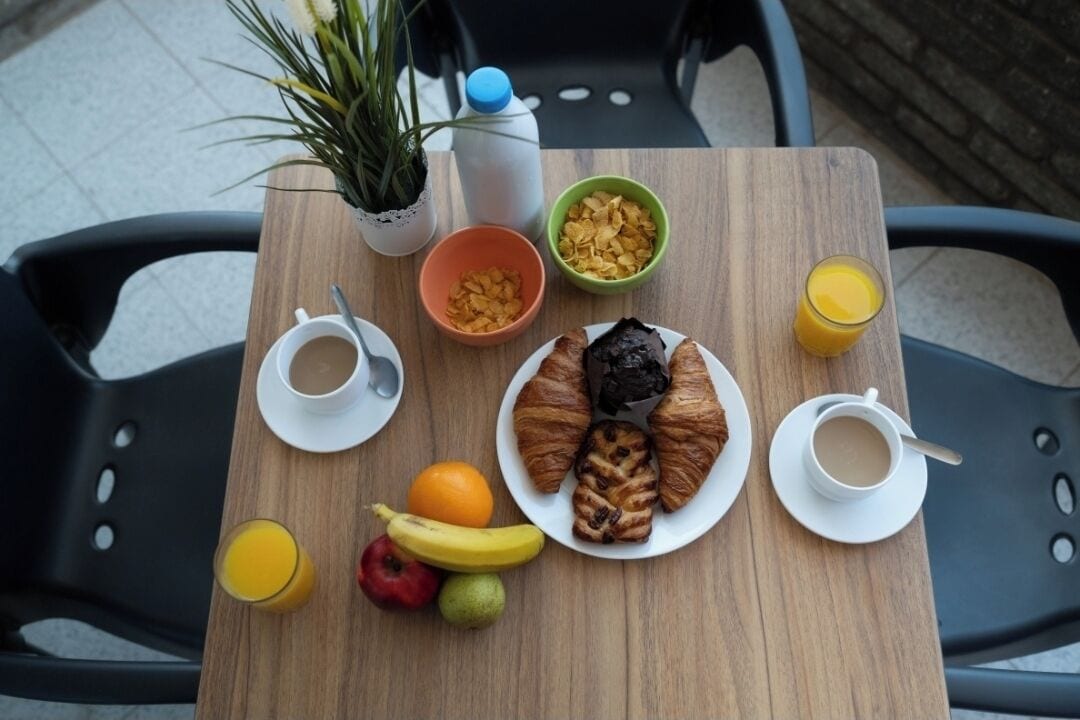 Ein Holztisch mit Frühstücksangeboten, darunter Croissants, Obst und Getränke, umgeben von schwarzen Stühlen.