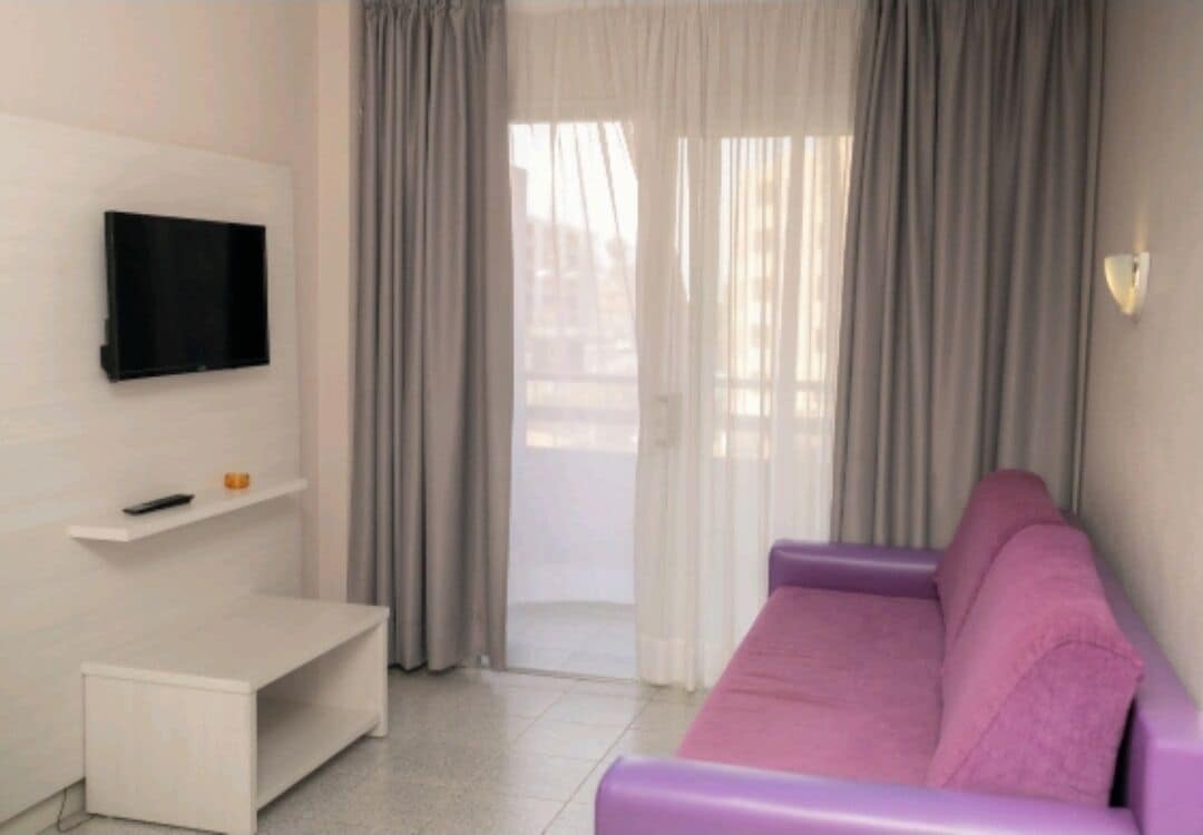A modern living room with a flat-screen TV mounted on the wall, a white bench, and a purple sofa.