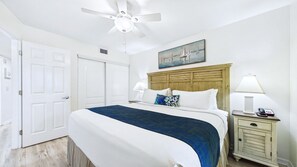 A hotel room with a bed, a nightstand, a lamp, a painting, a ceiling fan, and a door.