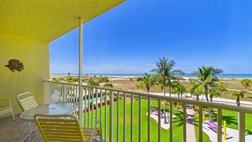 A balcony with a table and chairs, a view of the beach, and palm trees.