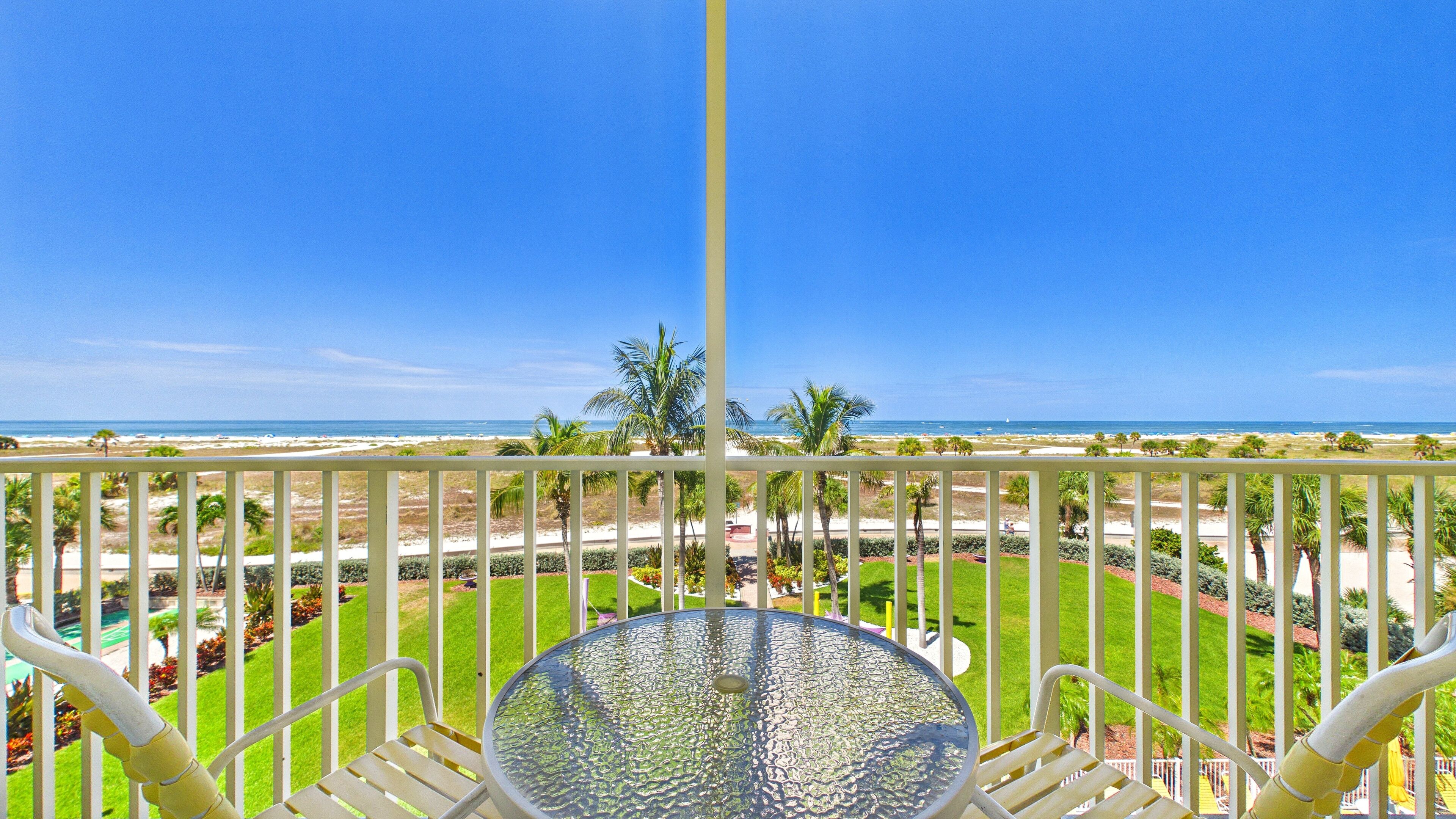 Un balcón con mesa y sillas, con vista a una playa con palmeras y un cielo despejado.