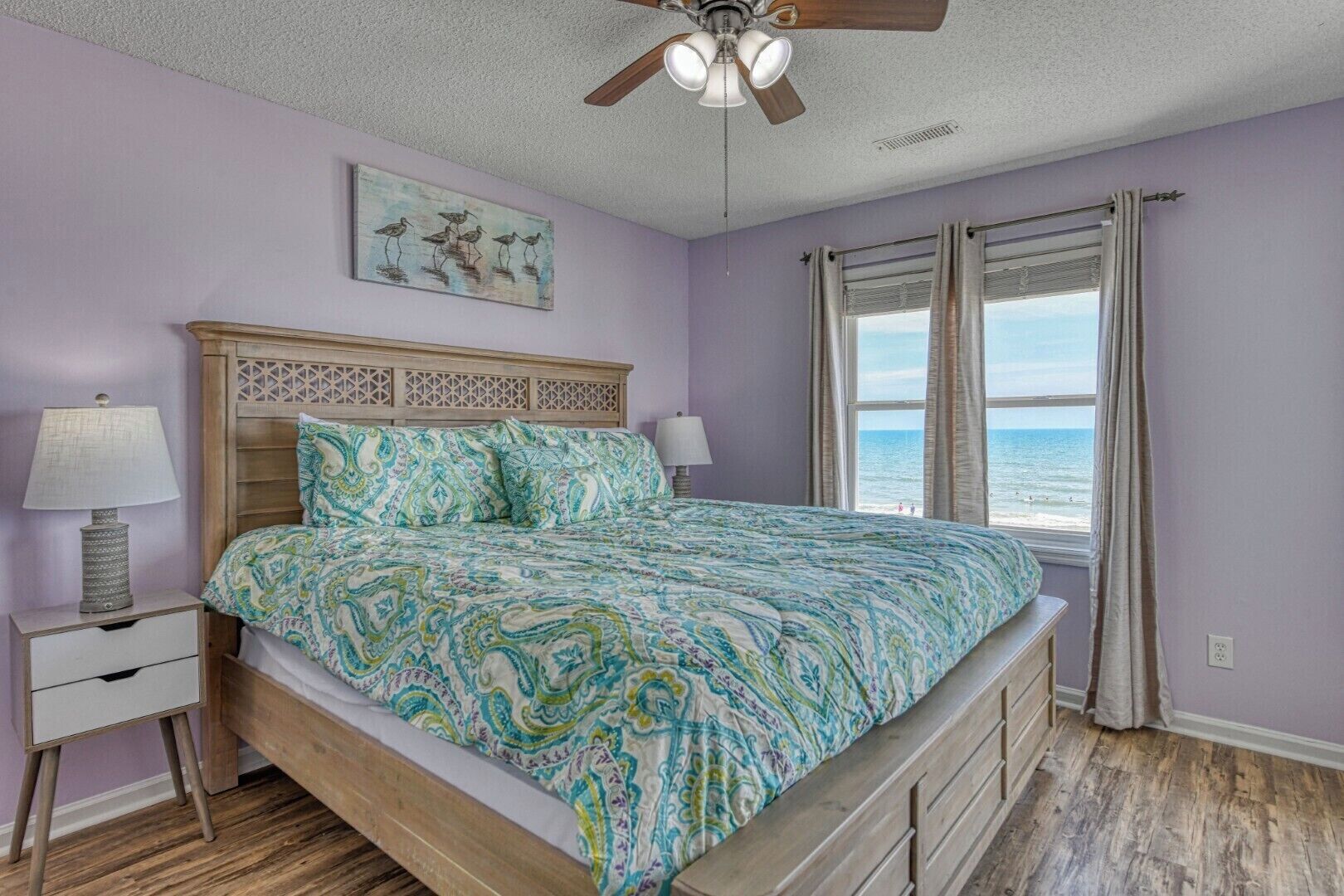 A bedroom with a bed, nightstands, a ceiling fan, a window with curtains, and a beach view.