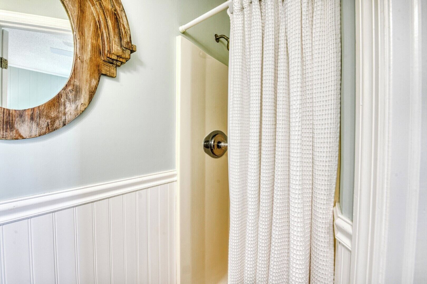 A bathroom with a wooden mirror, white walls, and a white door with a textured curtain.
