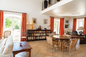 A spacious living room with a high ceiling, a dining area with a round table, and a library filled with books.
