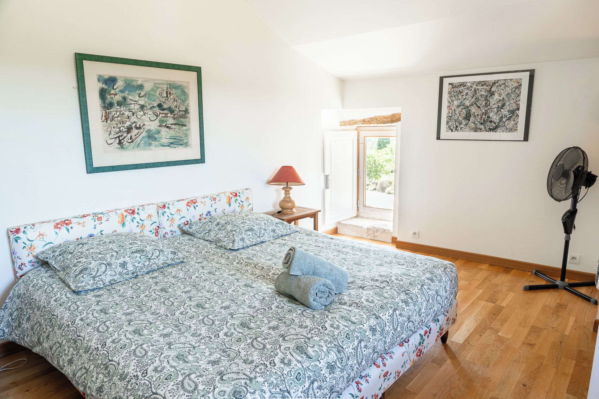A bedroom with a large bed, a standing fan, and two framed artworks on the walls.
