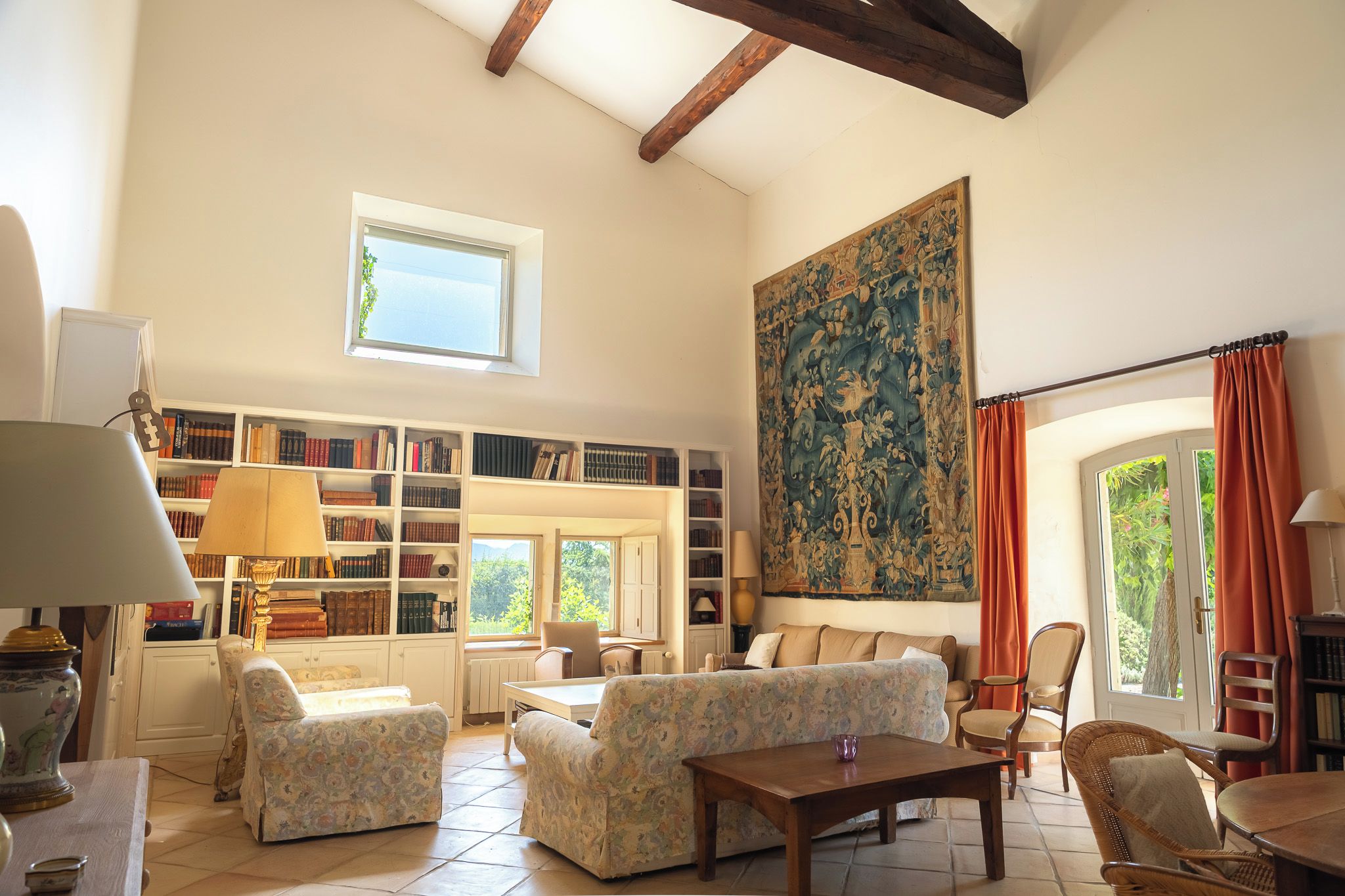 Een ruime woonkamer met een hoge nok, een grote boekenkast vol boeken, een groot wandtapijt en een gezellige zithoek met een bank en fauteuils.