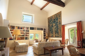 A spacious living room with a high ceiling, a large bookcase filled with books, a large tapestry, and a cozy seating area with a sofa and chairs.