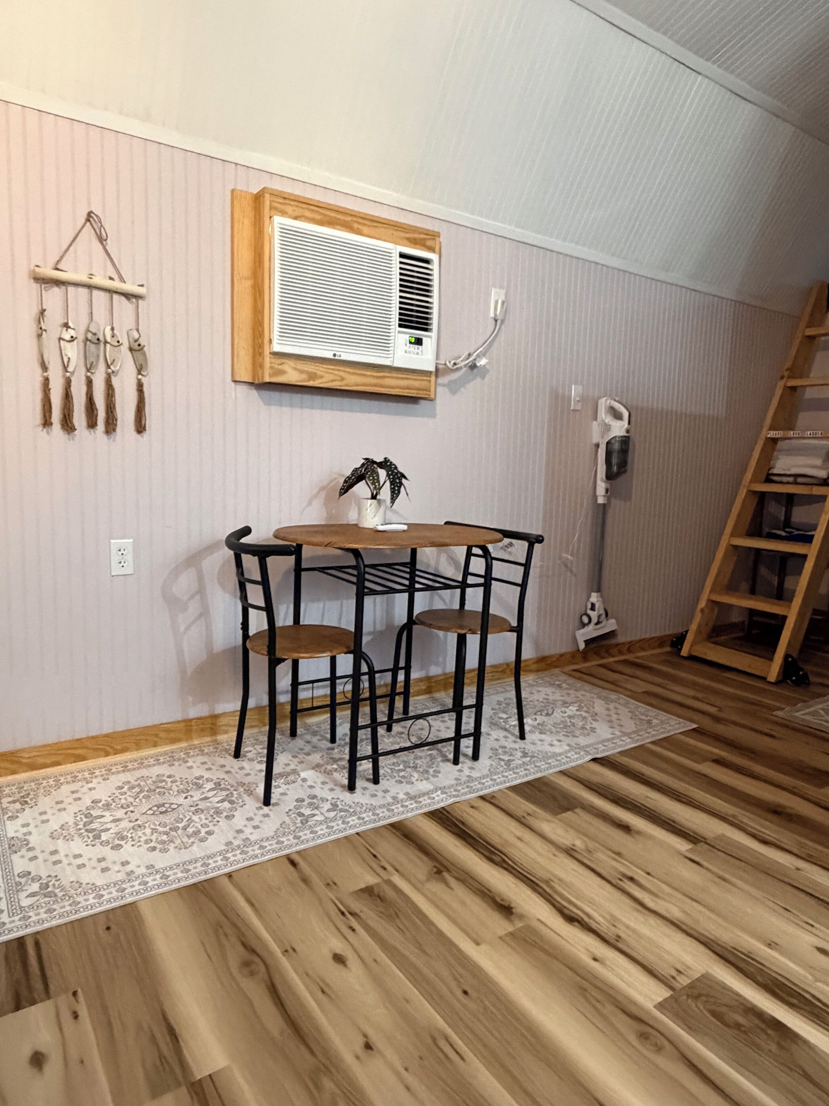 A room with a wooden floor, a table with chairs, a wall-mounted air conditioner, a ladder, and a hanging plant.