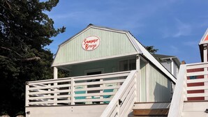 Eine Holztreppe führt zu einer überdachten Terrasse, auf der ein Schild mit der Aufschrift 'Summer Breeze' zu sehen ist, umgeben von viel Grün.