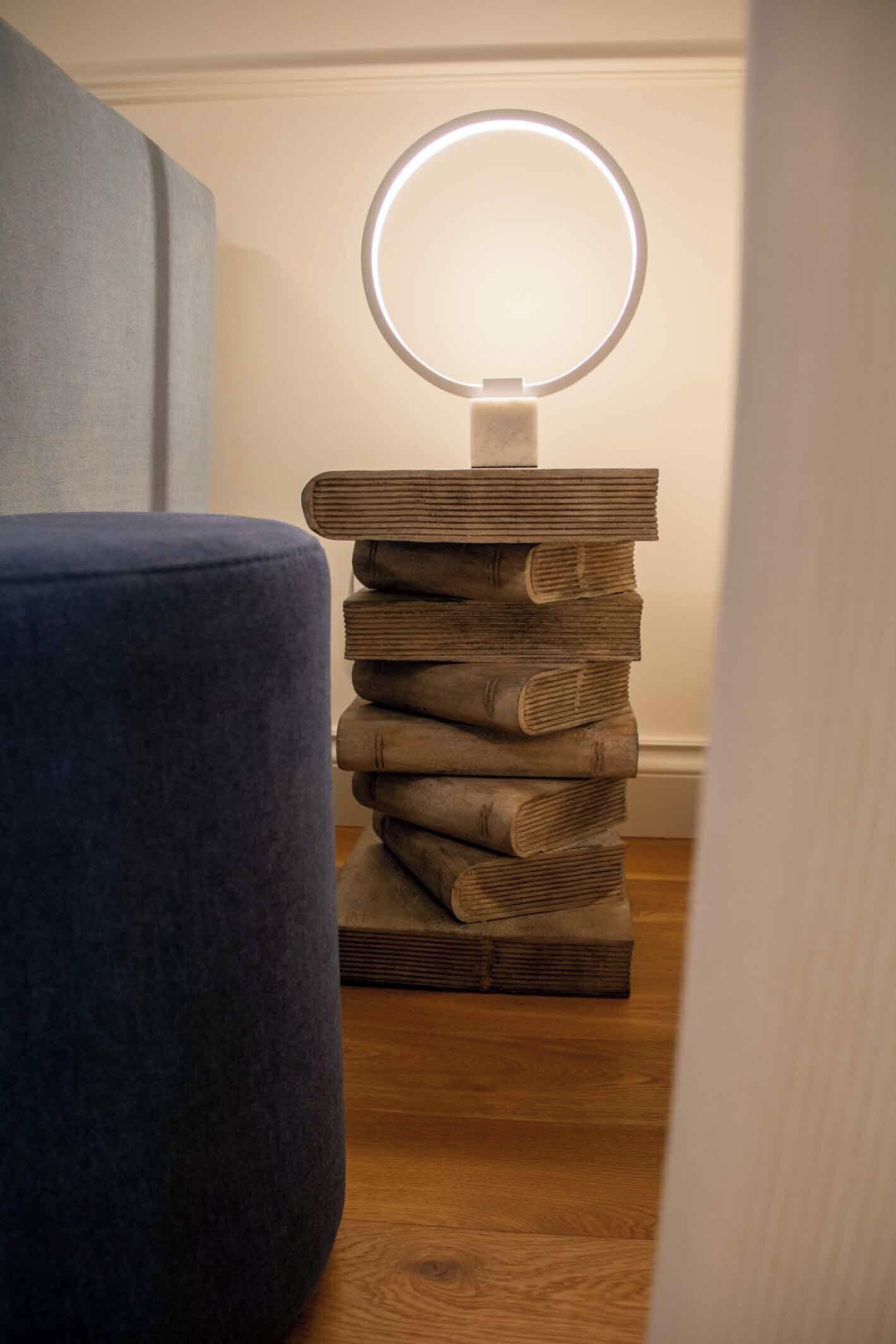 Una pila di libri con una luce circolare in cima, posizionata su un supporto in legno, accanto a una sedia blu.