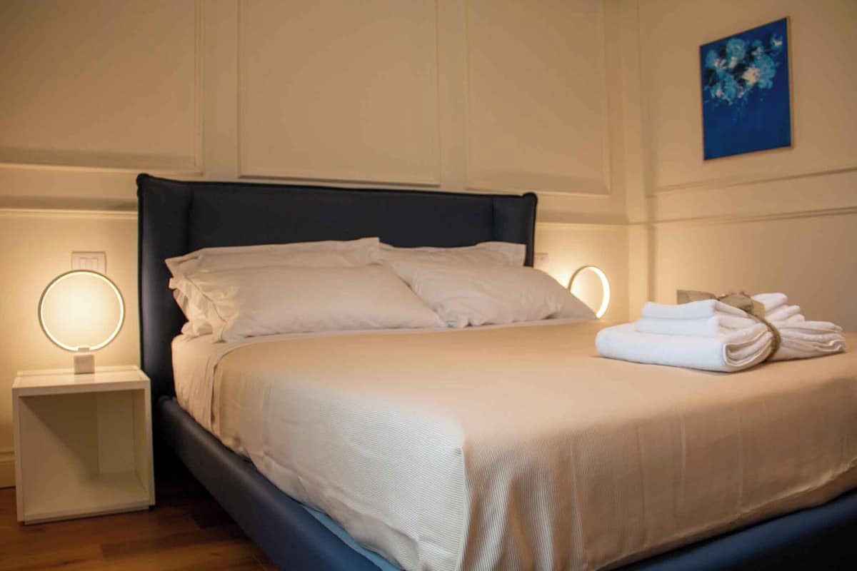 A neatly made bed with white linens, two bedside tables with illuminated lamps, and a blue abstract painting on the wall.