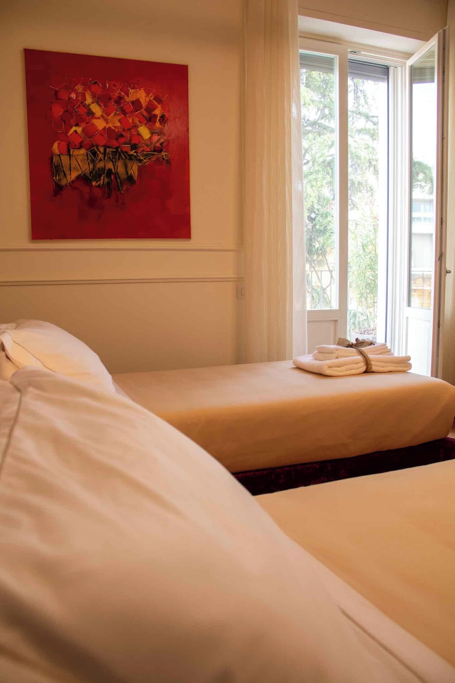Ein Hotelzimmer mit zwei Betten, einem roten abstrakten Gemälde an der Wand und einem Fenster mit Vorhängen.