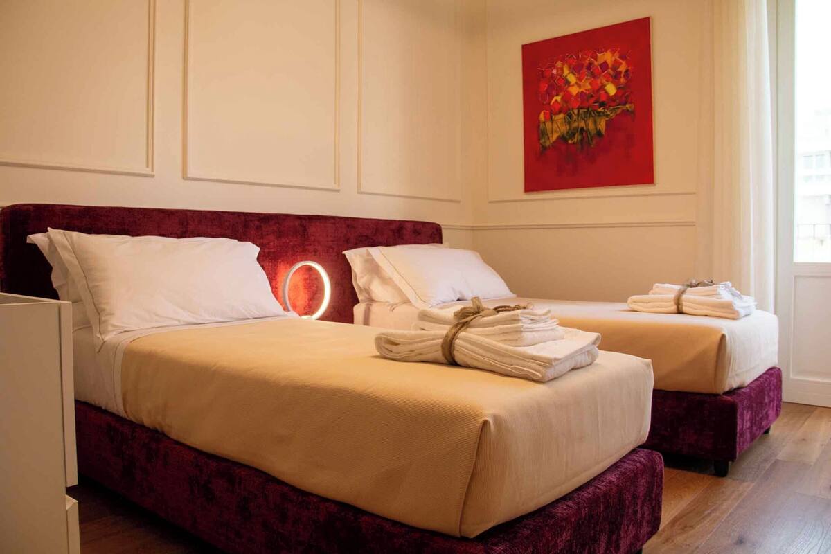 A hotel room with two beds, each with a red headboard and white bedding. There is a painting on the wall and a window with natural light.