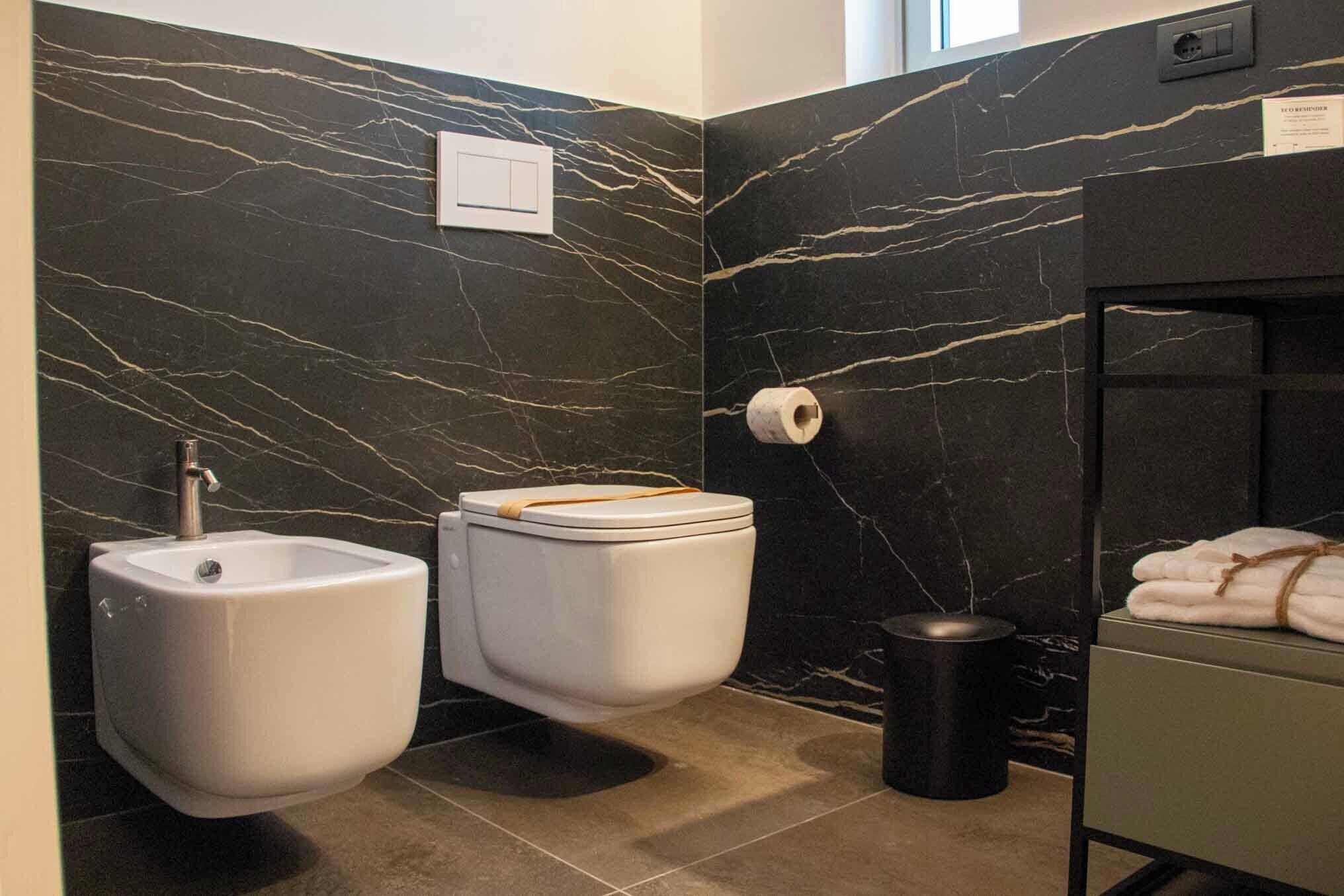 A modern bathroom with a bidet, toilet, marble walls, and a trash bin.