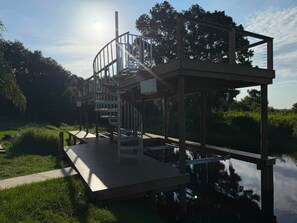 Una terrassa de fusta amb una escala que mena a una plataforma sobre l'aigua, envoltada de verdura.