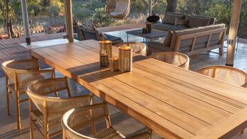 An outdoor patio with wooden tables and wicker chairs, a thatched roof, and a view of trees.