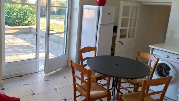Eine kleine Küche mit Esstisch und Stühlen, einem Kühlschrank, einer Waschmaschine und einem großen Fenster mit Blick ins Grüne.