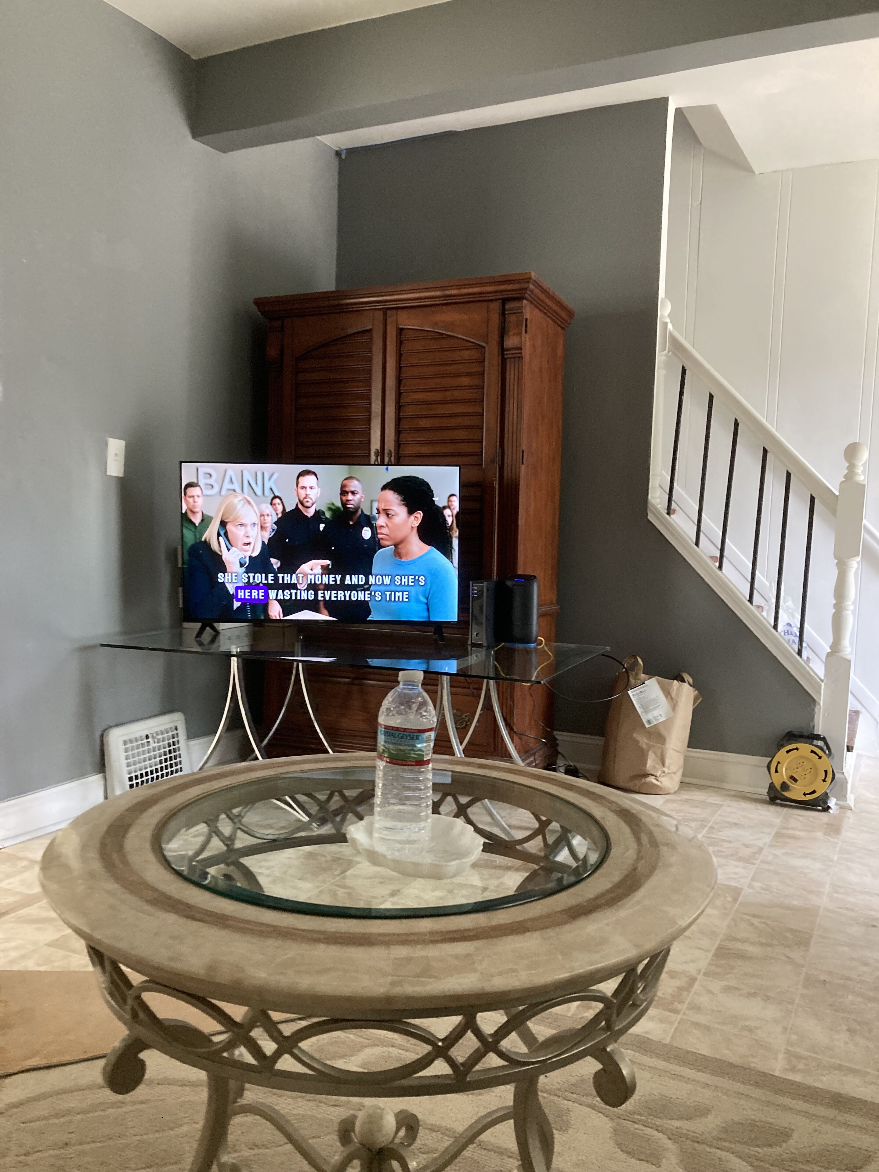A living room with a television displaying a show, a wooden cabinet, a glass coffee table, and a staircase.