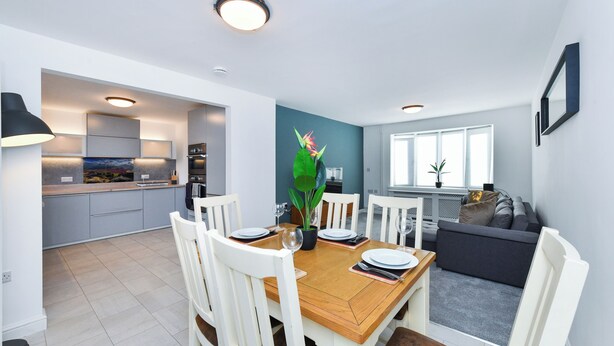 Una cocina moderna con armarios blancos, una mesa de comedor preparada para cuatro personas y un sofá gris en la sala.
