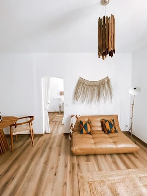 A room with a wooden floor, a brown leather sofa with patterned cushions, a hanging wooden decor, and a white lamp. - Casa SaloYa (Sintra)