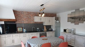 A kitchen with white cabinets, a brick wall, a dining table with a patterned tablecloth, and red chairs. - Stone house 600 m from the sea with garden and terrace. (Plomodiern)