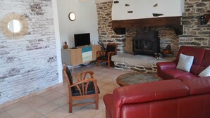 A living room with a stone fireplace, a red sofa, a wooden chair, a TV on a wooden cabinet, and a circular light fixture. - Stone house 600 m from the sea with garden and terrace. (Plomodiern)