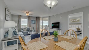 A modern living room with a dining area, grey walls, and a large window. - 3BR- 12th floor- Lake & Golf View condo|Short walk to beach| Resort Access| (North Myrtle Beach)