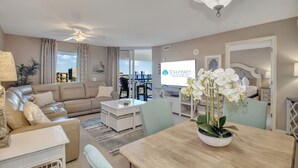 Un salon spacieux avec un canapé beige, une table à manger agrémentée d’un vase de fleurs blanches, et une vue sur la ville grâce à de grandes fenêtres.