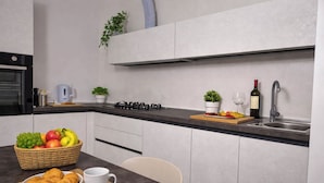 A modern kitchen with a built-in stove, a sink, and a countertop. There are two potted plants, a basket of fruit, and a bowl of croissants on the counter.