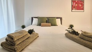 A neatly made bed with a white duvet and two pillows, a small potted plant on the left side, and a framed artwork on the wall.