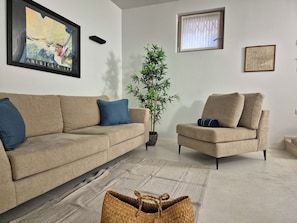 Un salon avec un canapé beige, un coussin bleu, une petite table d’appoint, un panier en osier, une plante en pot et une carte encadrée accrochée au mur.