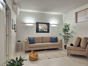 Un salon avec un canapé beige, un coussin bleu, un panier en osier et une grande plante en pot.