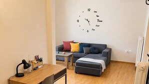 A compact living space with a wooden floor, a desk with a chair, a sofa, and a wall clock. - Alimos Cozy Getaway, close to the beach (Alimos)