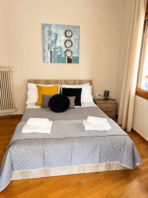 A neatly made bed with a grey quilt, white pillows, and a black round pillow. There is a painting above the bed, a nightstand with a lamp, and a window with curtains.